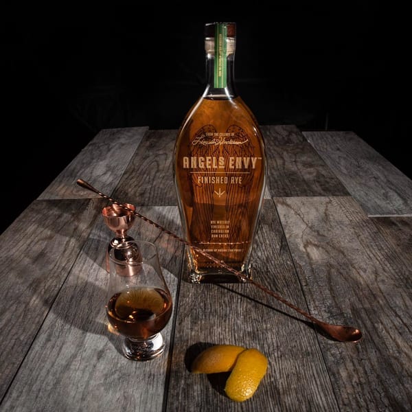 Angel's Envy Finished Rye bottle, glass of whiskey, jigger, bar spoon, and lemon peels on wood.