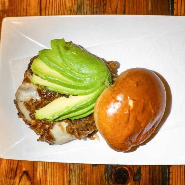 An open-faced burger with caramelized onions, melted cheese, and sliced avocado on a bun.