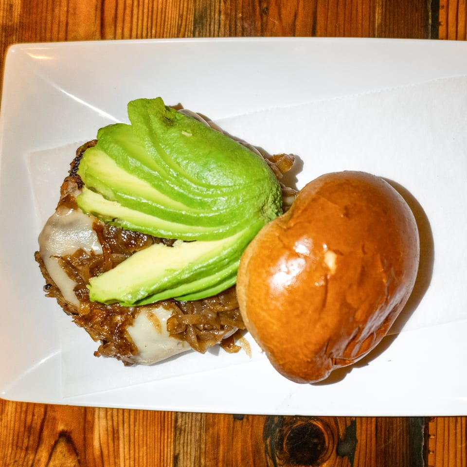 An open-faced burger with caramelized onions, melted cheese, and sliced avocado on a bun.