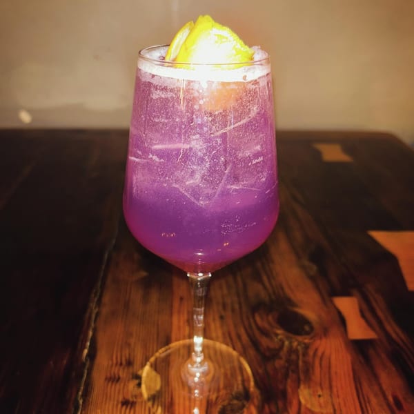 A vibrant purple cocktail with lemon slices and ice sits on a dark wooden table.