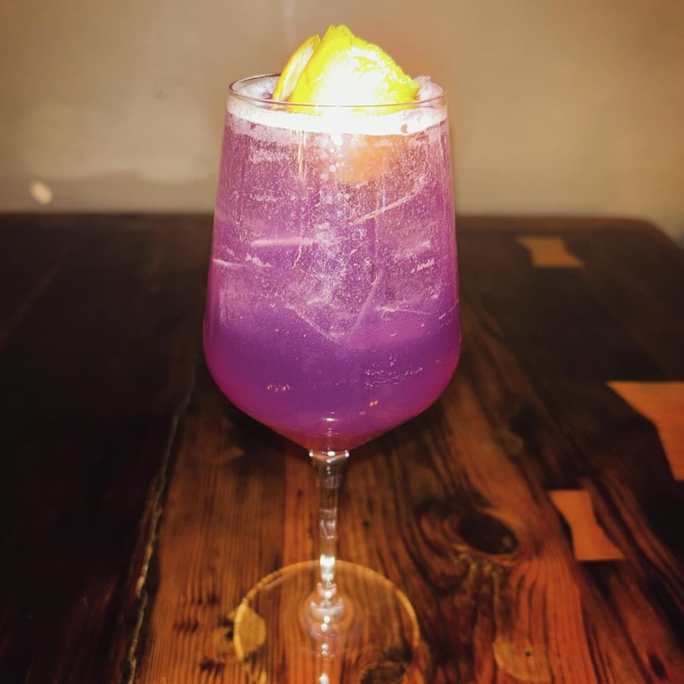 A vibrant purple cocktail with lemon slices and ice sits on a dark wooden table.