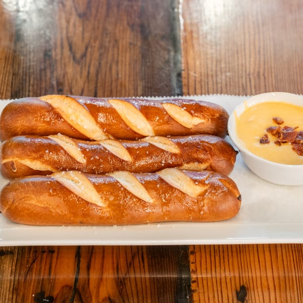 Three salted pretzel sticks and a bowl of cheese dip with bacon bits on a white plate.