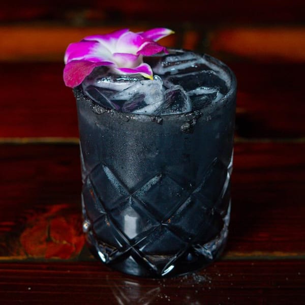 Dark cocktail with ice and a purple orchid garnish in a cut glass on a wooden surface.