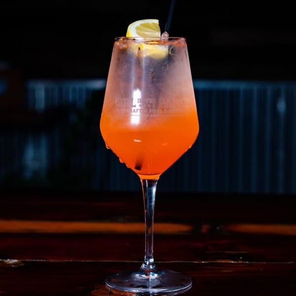A Girl in the Park branded glass filled with an orange cocktail, garnished with a lemon slice.