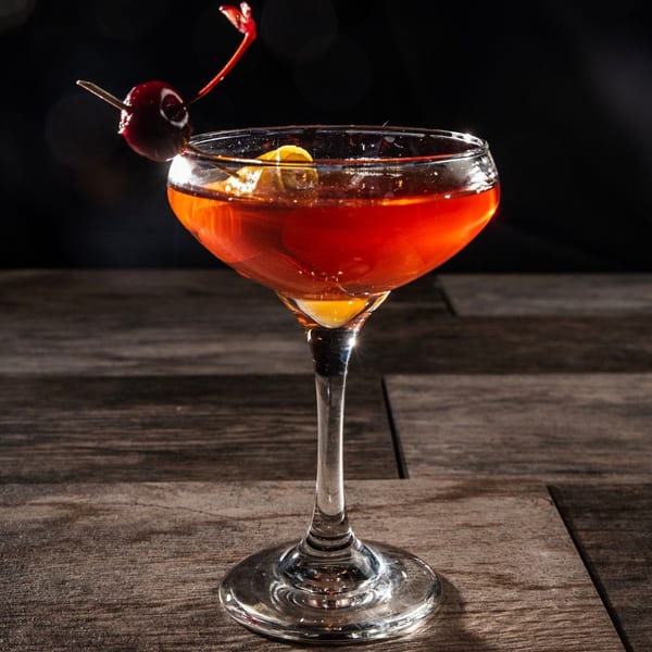 A red cocktail in a coupe glass, garnished with a cherry and lemon peel, on a wooden table.